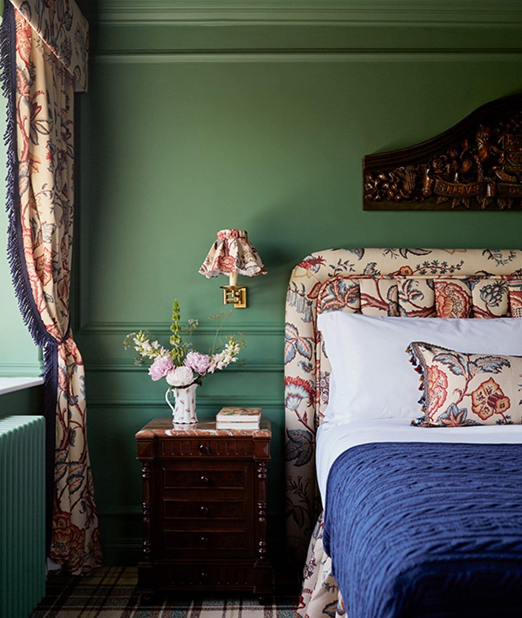 A bedroom with a green wall, floral patterned bedspread, a bedside table with a vase of flowers, and a lamp.