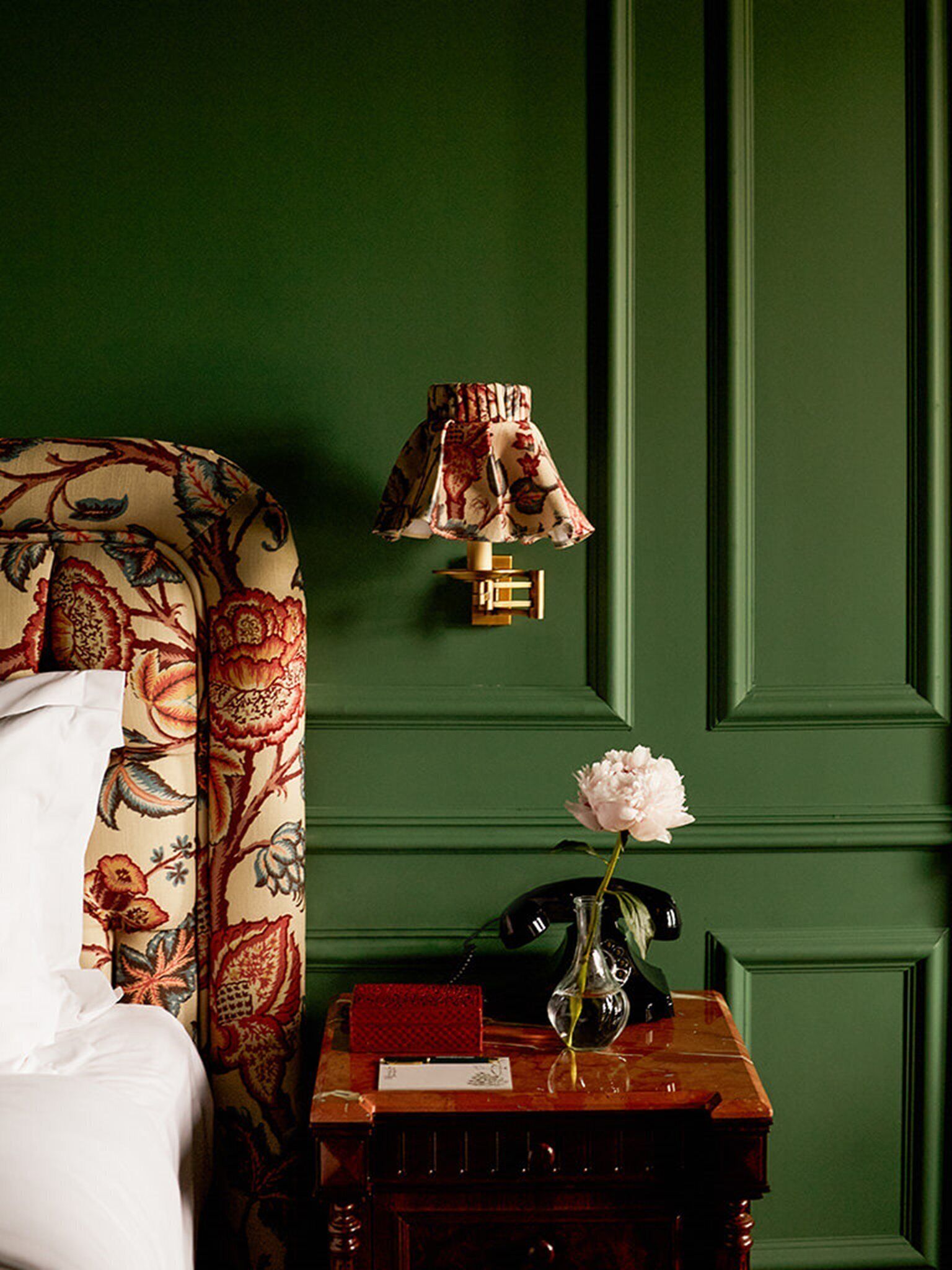 A room with a green wall, a patterned headboard, a bedside table with a vase of flowers, a telephone, and a red book.