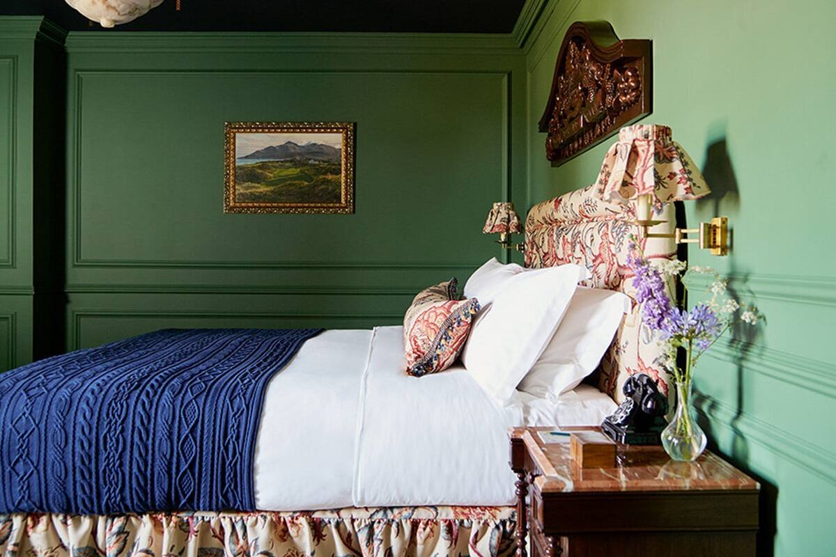 A neatly made bed with a blue blanket, a floral-patterned pillow, and a white comforter. A wooden nightstand with a vase of flowers and a lamp is beside the bed.