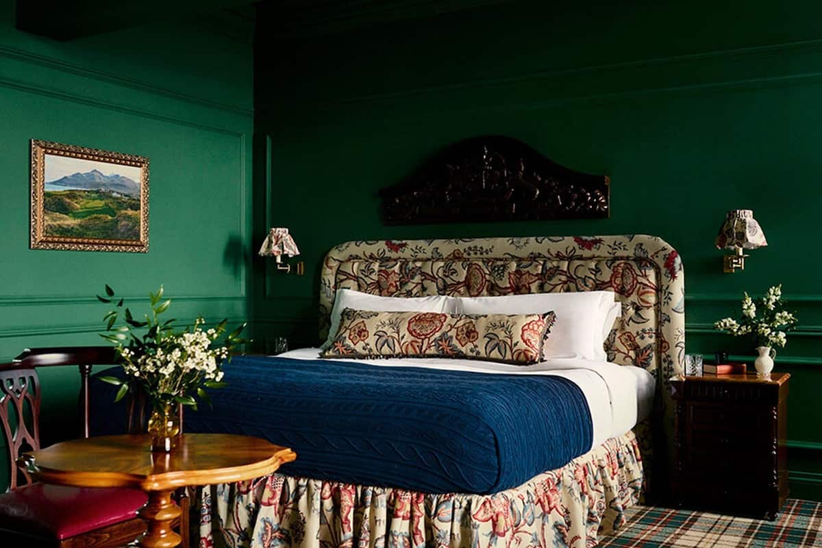A room with a green wall, a bed with floral bedding, a wooden chair, a small table with a vase, and a framed picture on the wall.
