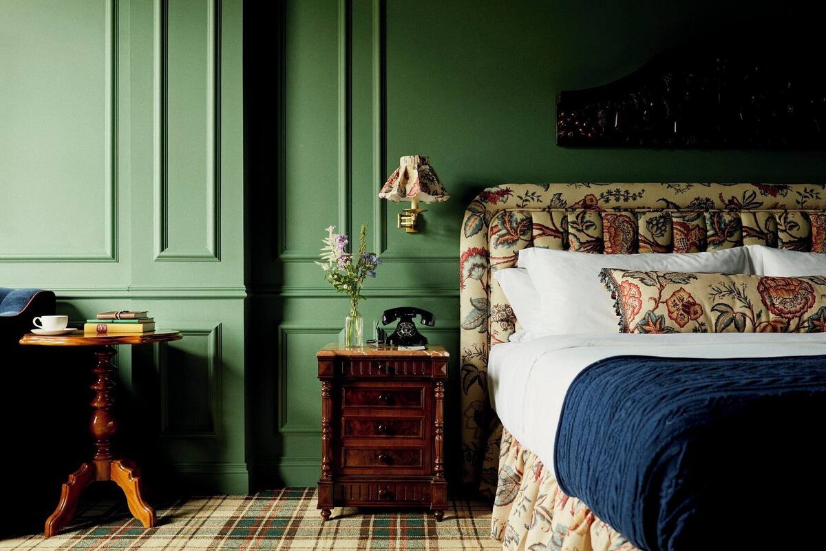 A room with a green wall, a patterned bed, a small wooden nightstand with drawers, a vintage telephone, and a vase with flowers.