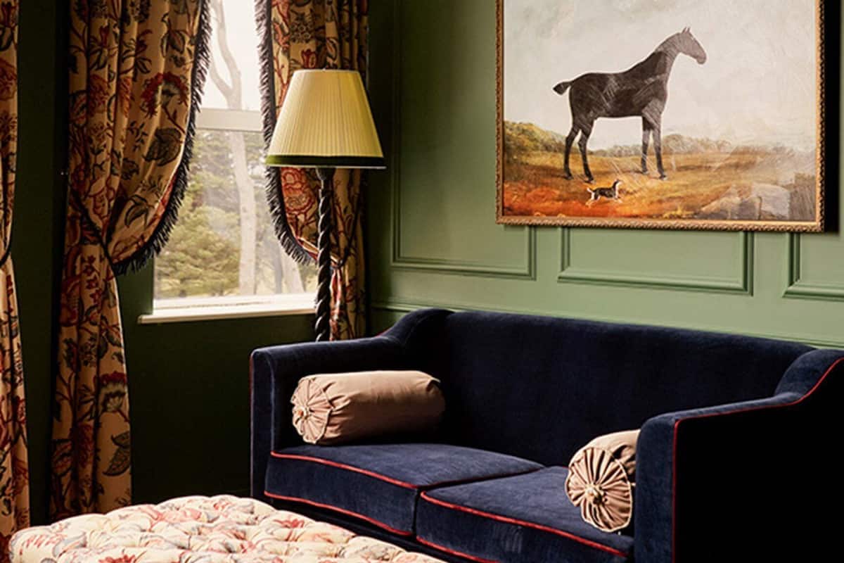 A room with a green wall, a blue sofa with red trim, a patterned ottoman, a lamp, and a painting of a horse.