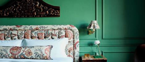 Una cama con ropa de cama floral, cabecera de madera, mesita de noche con lámpara y pared verde.