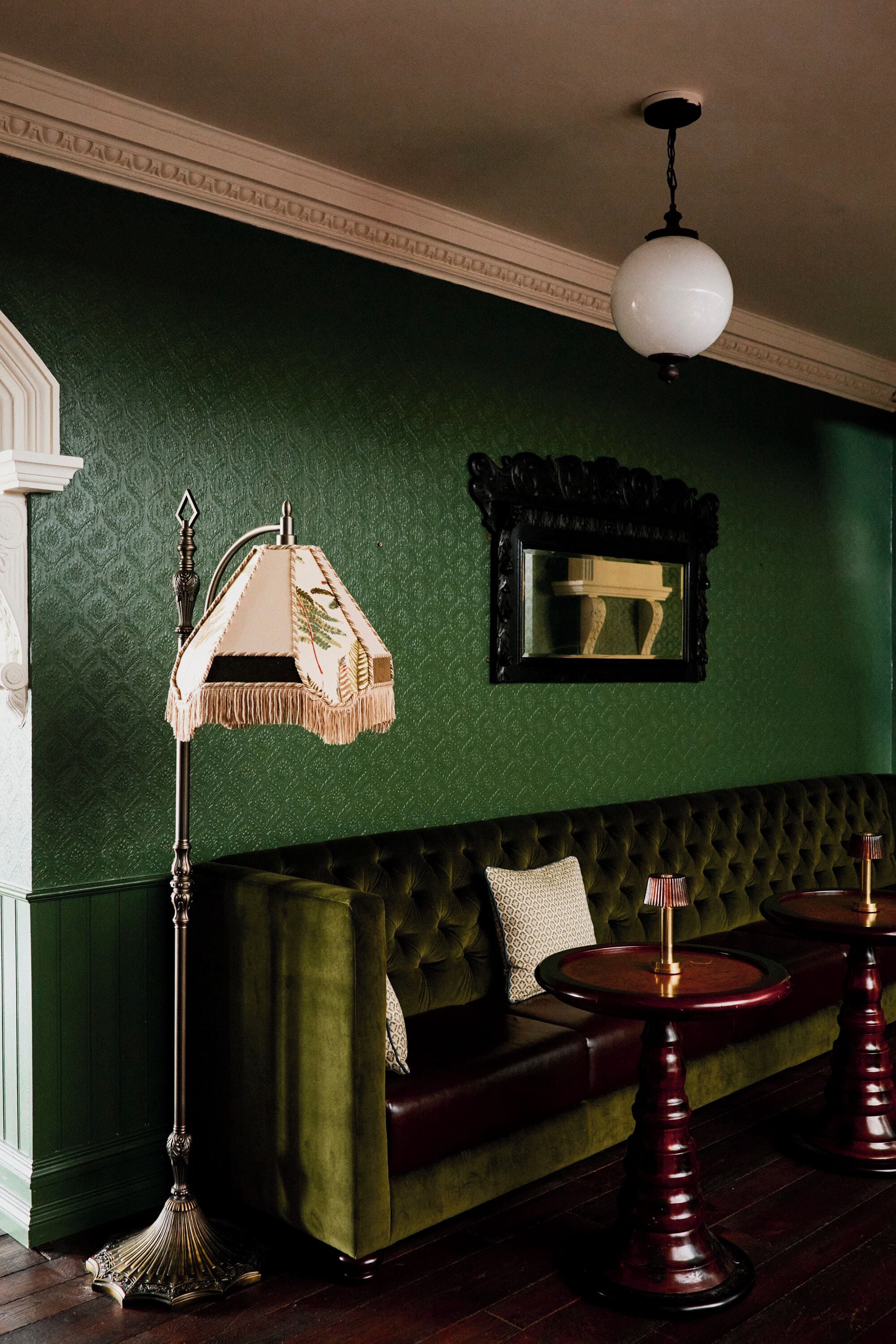A green upholstered sofa with a mirror above it, a floor lamp, and two round wooden tables.