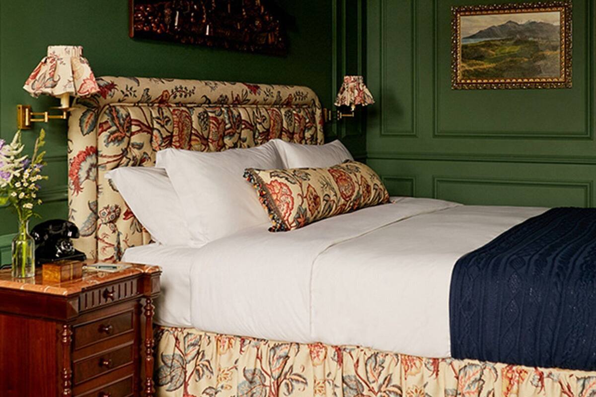 A bedroom with a floral-patterned bedspread, green walls, a wooden nightstand, and a framed landscape painting.