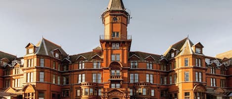 Un edificio grande e histórico con una torre destacada y varias secciones. Cuenta con una entrada imponente con escalones y plantas en macetas.
