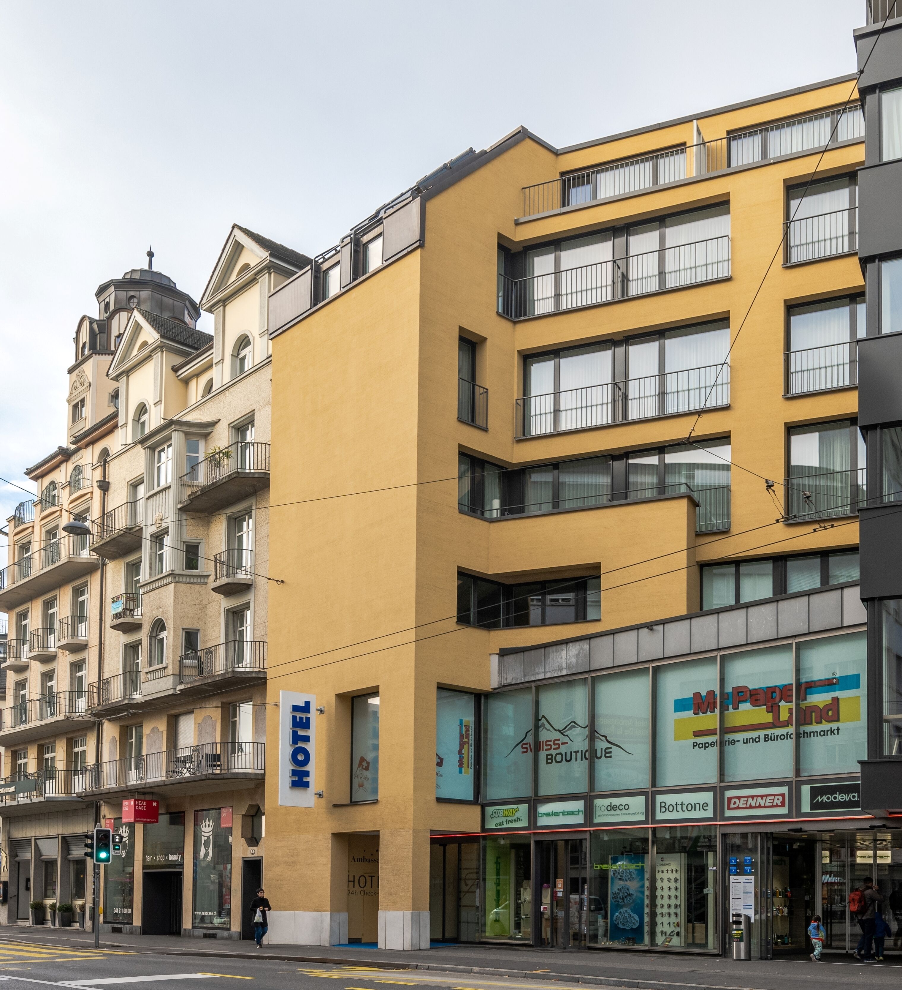 Um hotel com fachada amarela, vários andares e diversos estabelecimentos comerciais no térreo.