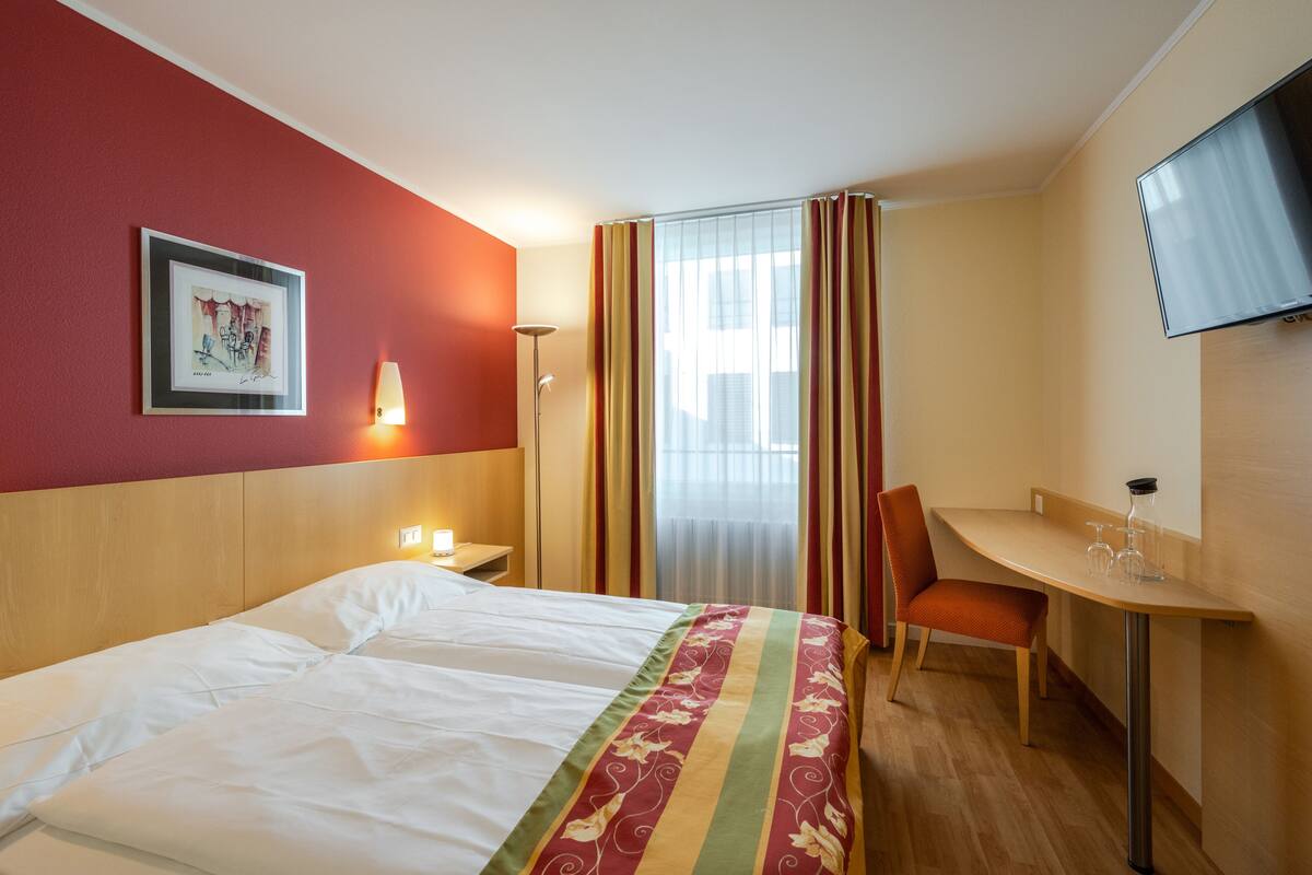 Una habitación de hotel con cama, escritorio, silla, televisión y ventana con cortinas.