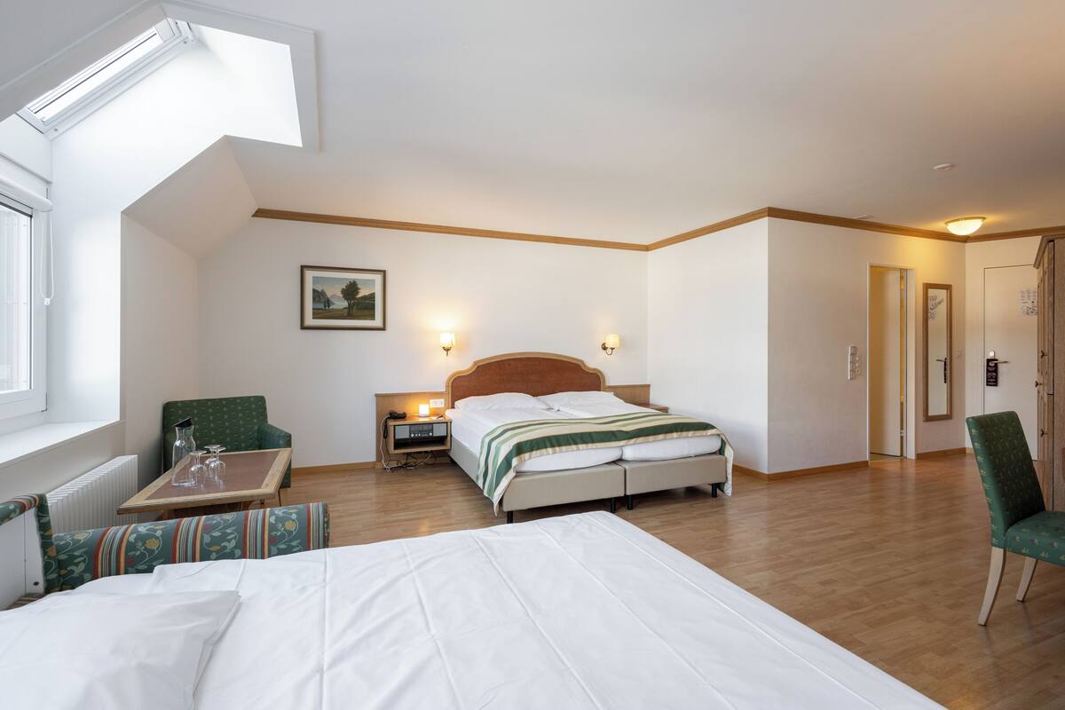 Una habitación de hotel con cama, una silla, una mesa, una lámpara de noche, un cuadro en la pared y una ventana.