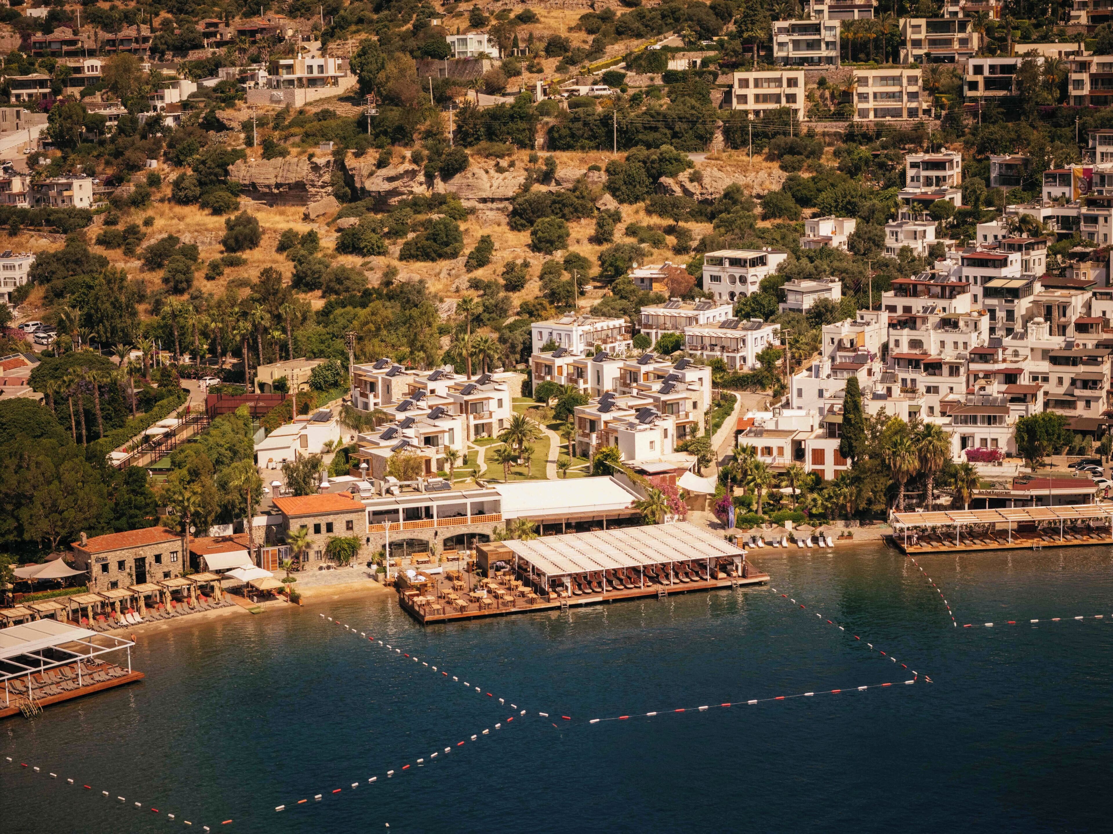 A coastal town with modern buildings, a marina with boats, and a restaurant with outdoor seating.