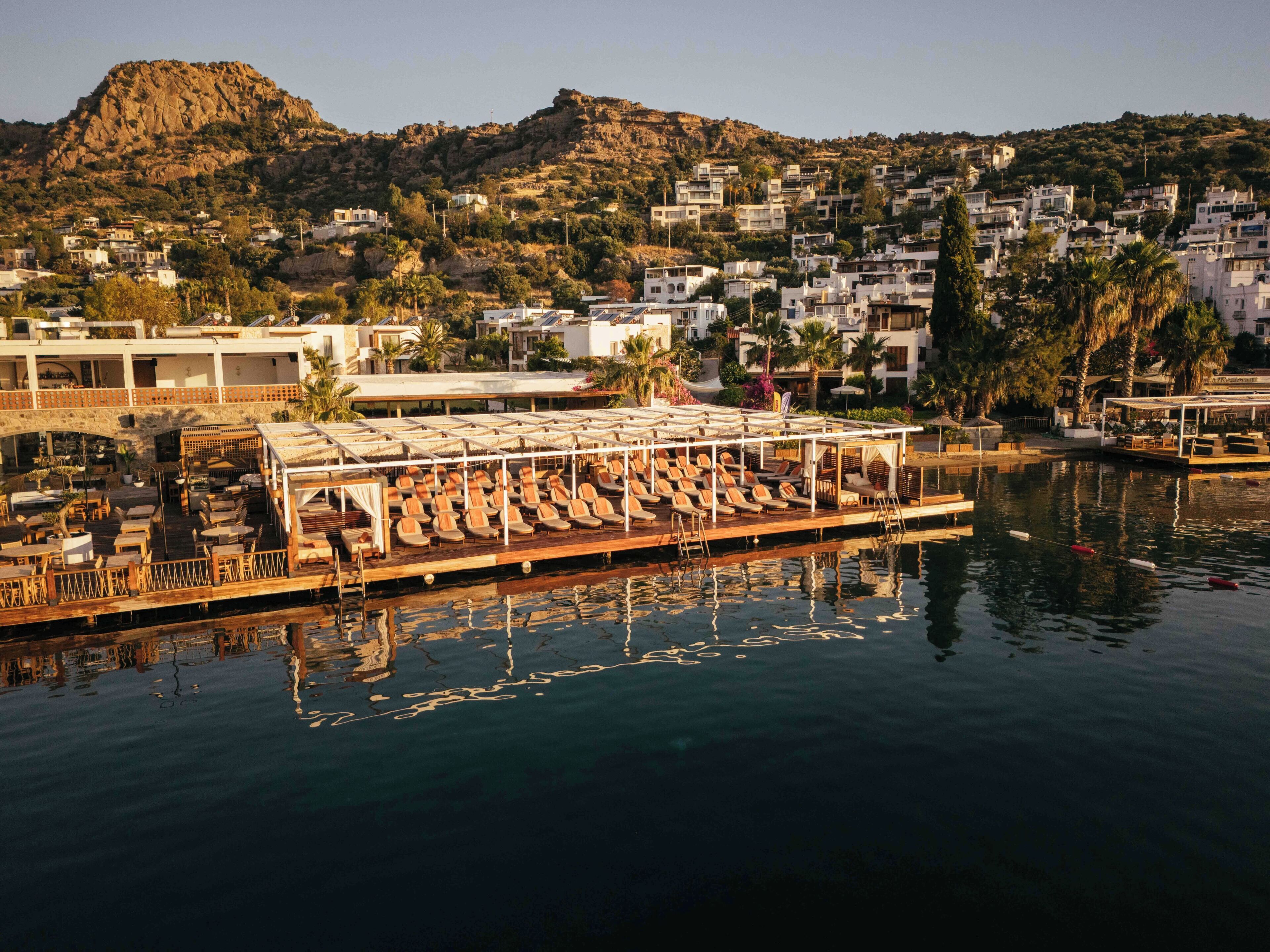 A waterfront hotel with a poolside area, a mountain backdrop, and a view of the town.