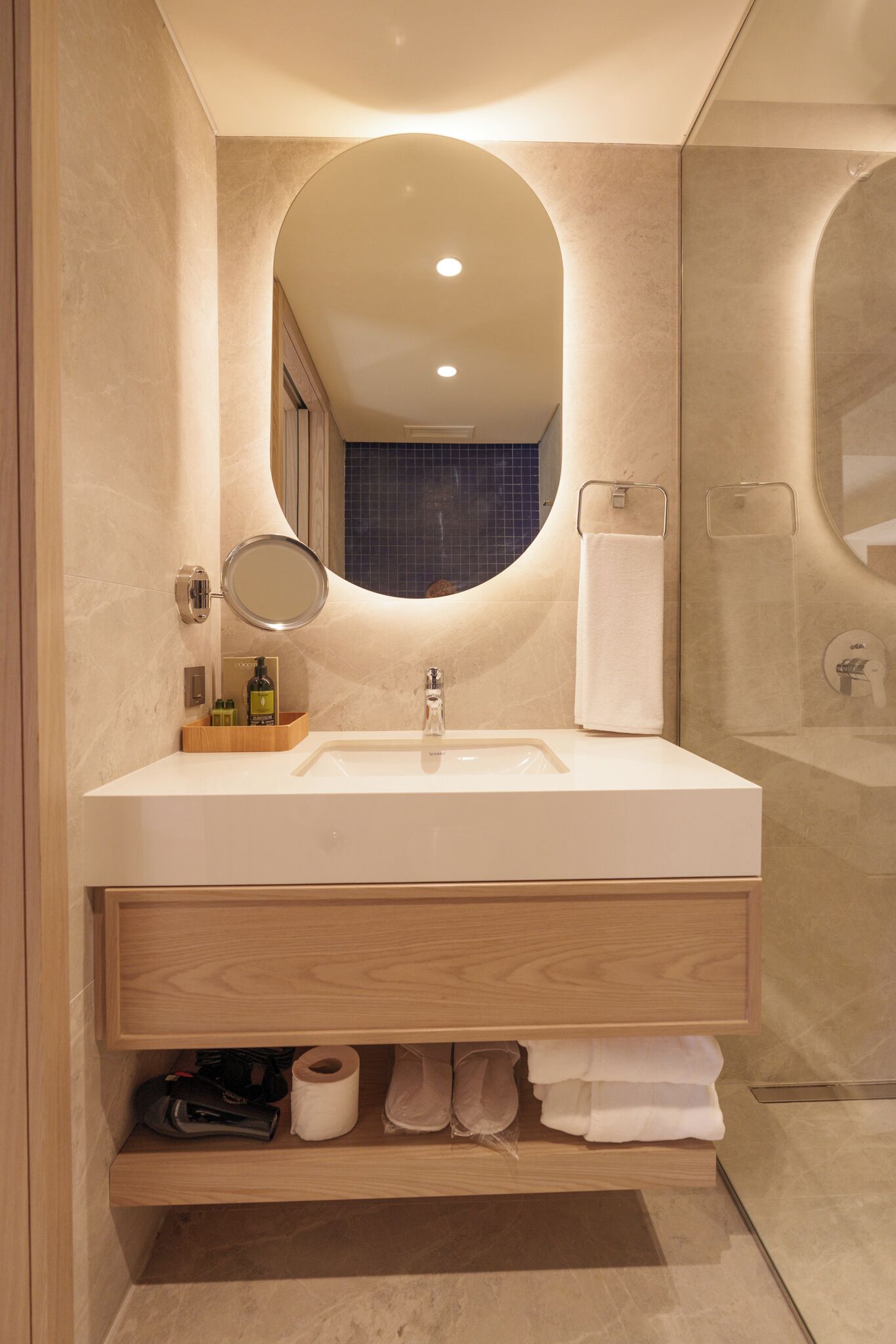 A modern bathroom with a large oval mirror, a white sink, and a wooden vanity.