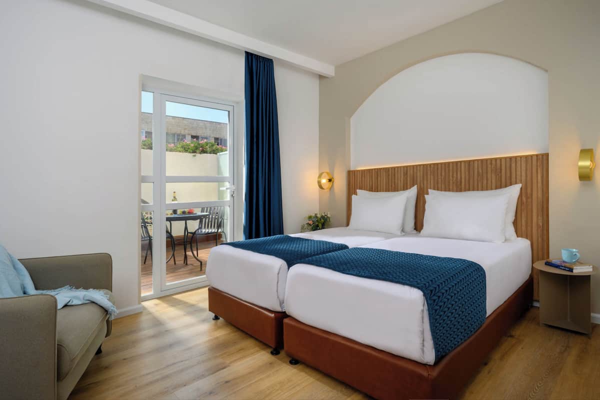 Une chambre d’hôtel avec un lit double, un canapé, une petite table avec une tasse et un livre, et une vue sur le balcon extérieur.