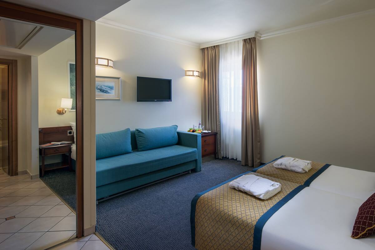 Une chambre d’hôtel avec un lit, un canapé bleu, une télévision, une table de chevet et une fenêtre avec des rideaux.