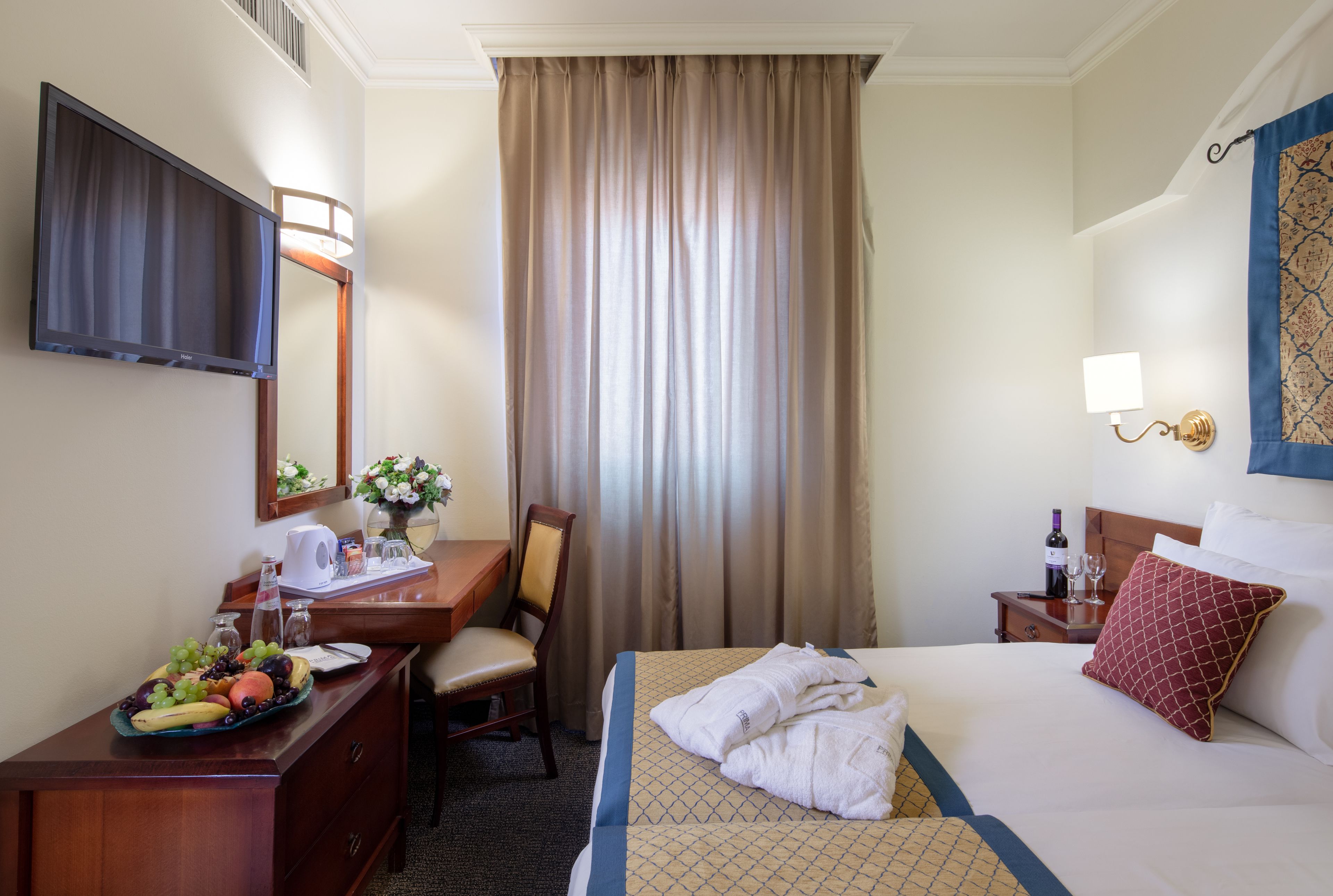 A hotel room with a bed, a desk with a fruit bowl, a chair, a TV, a nightstand with a bottle and glasses, and a window with curtains.