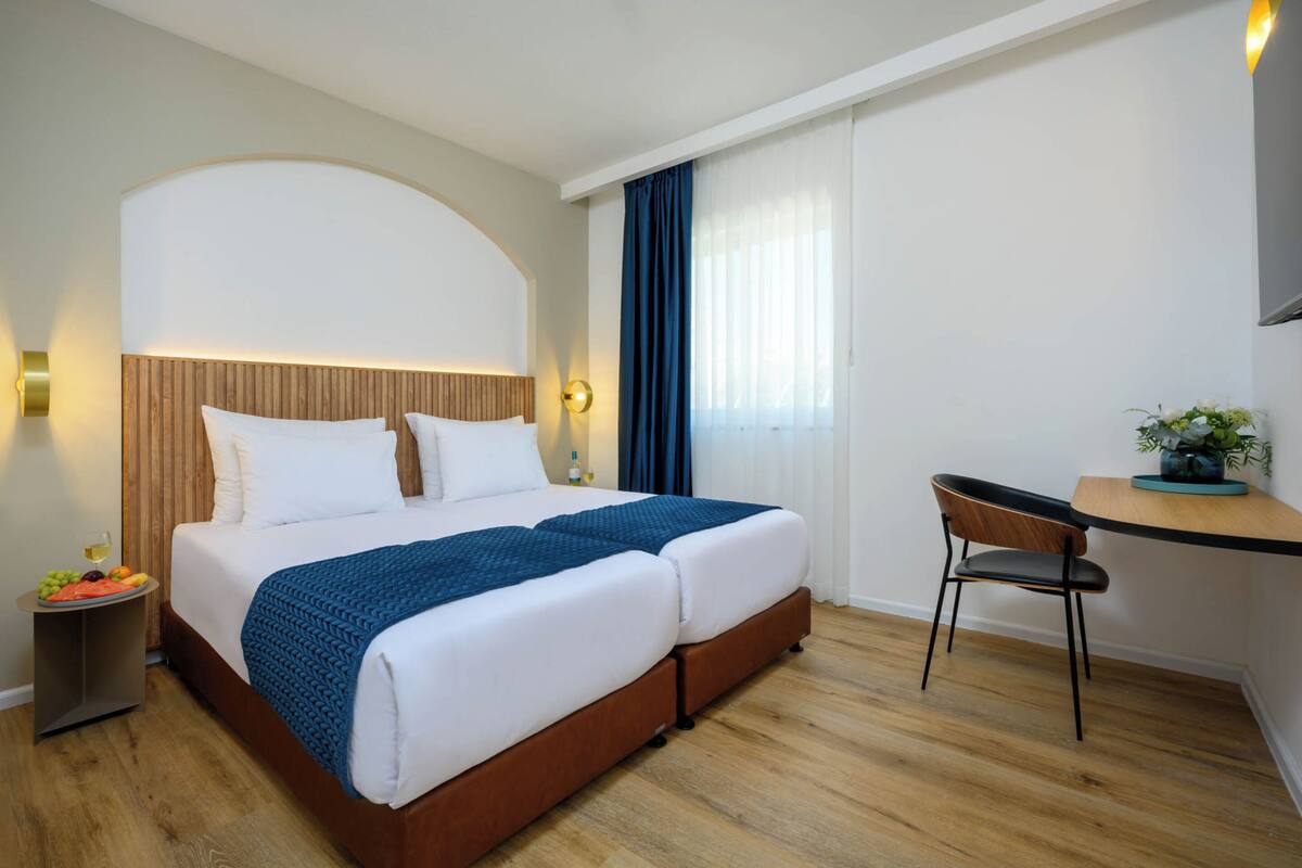 Une chambre d’hôtel avec un lit double, un bureau avec une chaise, une petite table d’appoint avec des fruits et une plante en pot.