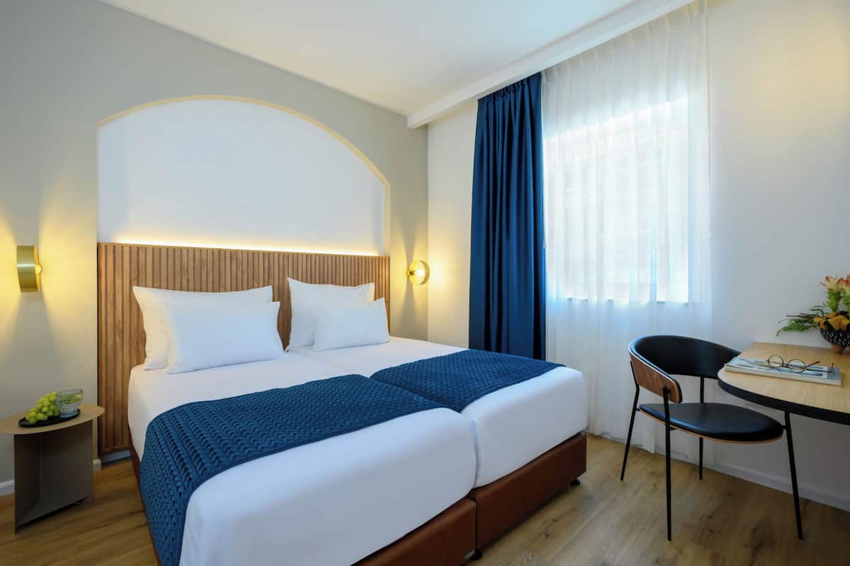 Une chambre d’hôtel avec un grand lit, un bureau avec une chaise, une petite table avec un panier de fruits et une fenêtre avec des rideaux.