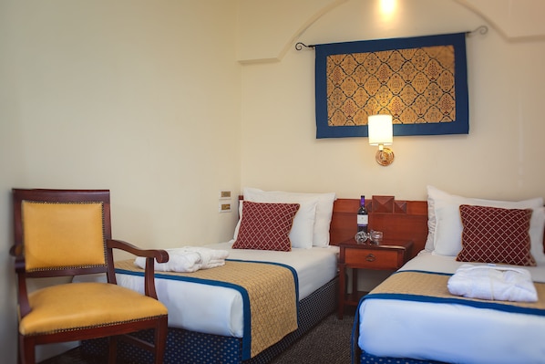 A hotel room with two beds, a wooden chair, a nightstand, and a wall-mounted lamp.