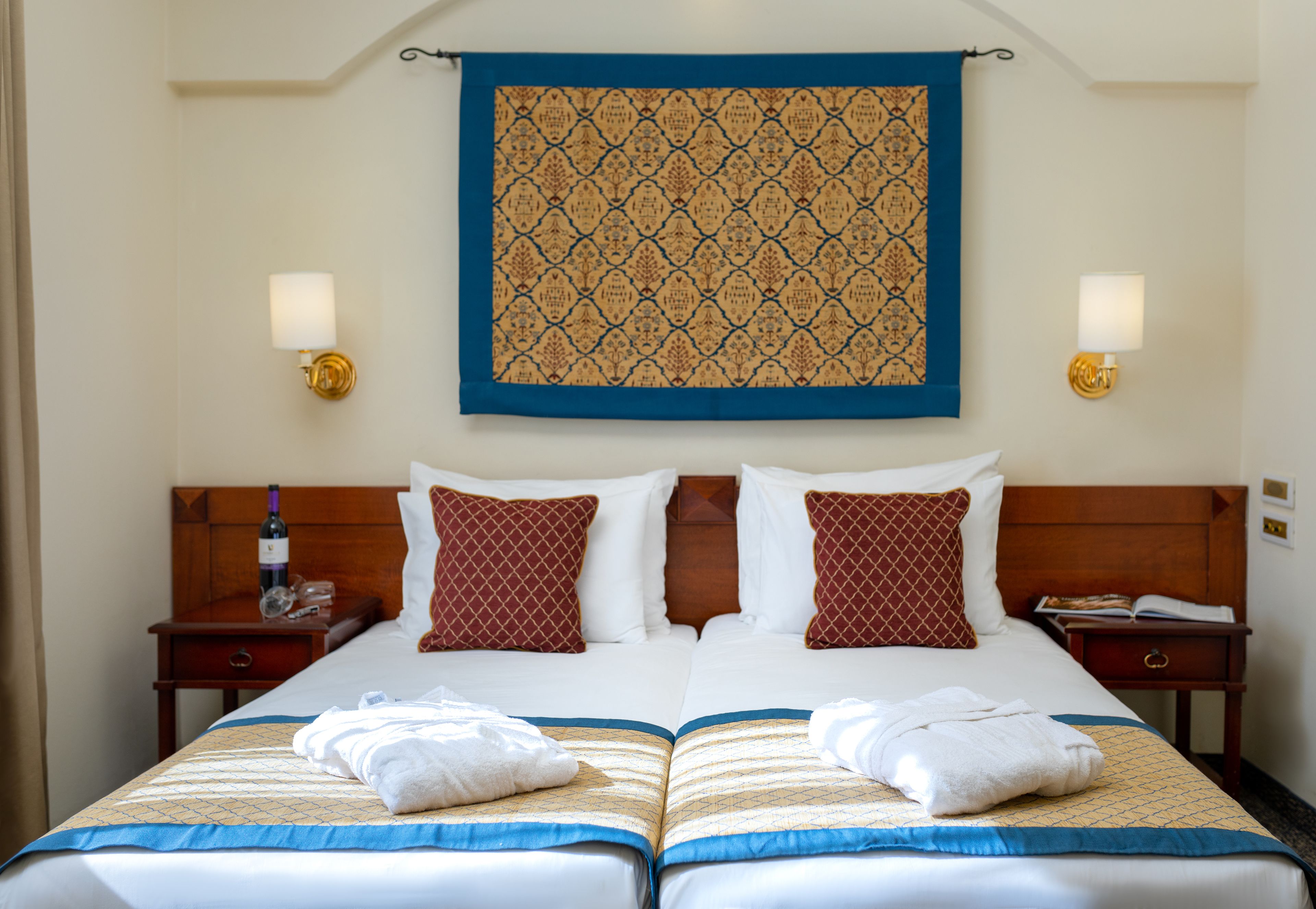 A double bed with a blue and gold bedspread, two pillows, a wooden headboard, and a wall tapestry.