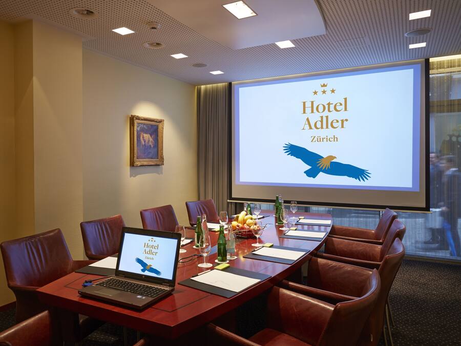A conference room with a large screen displaying "Hotel Adler Zurich" and a laptop on the table.