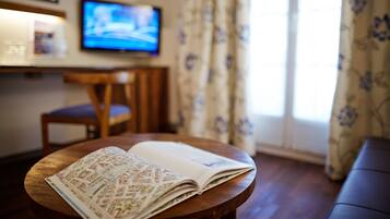 Quarto com mesa de madeira, revista aberta sobre ela, televisão na parede e janela com cortinas.
