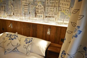 A bedroom with a wooden headboard, a bed with white bedding and blue floral pillows, a nightstand, and a window with a cityscape view.