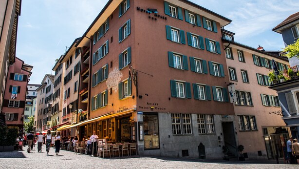 Et hotel med flere etager, restaurant i stueplan, flere balkoner og en brostensbelagt gade.