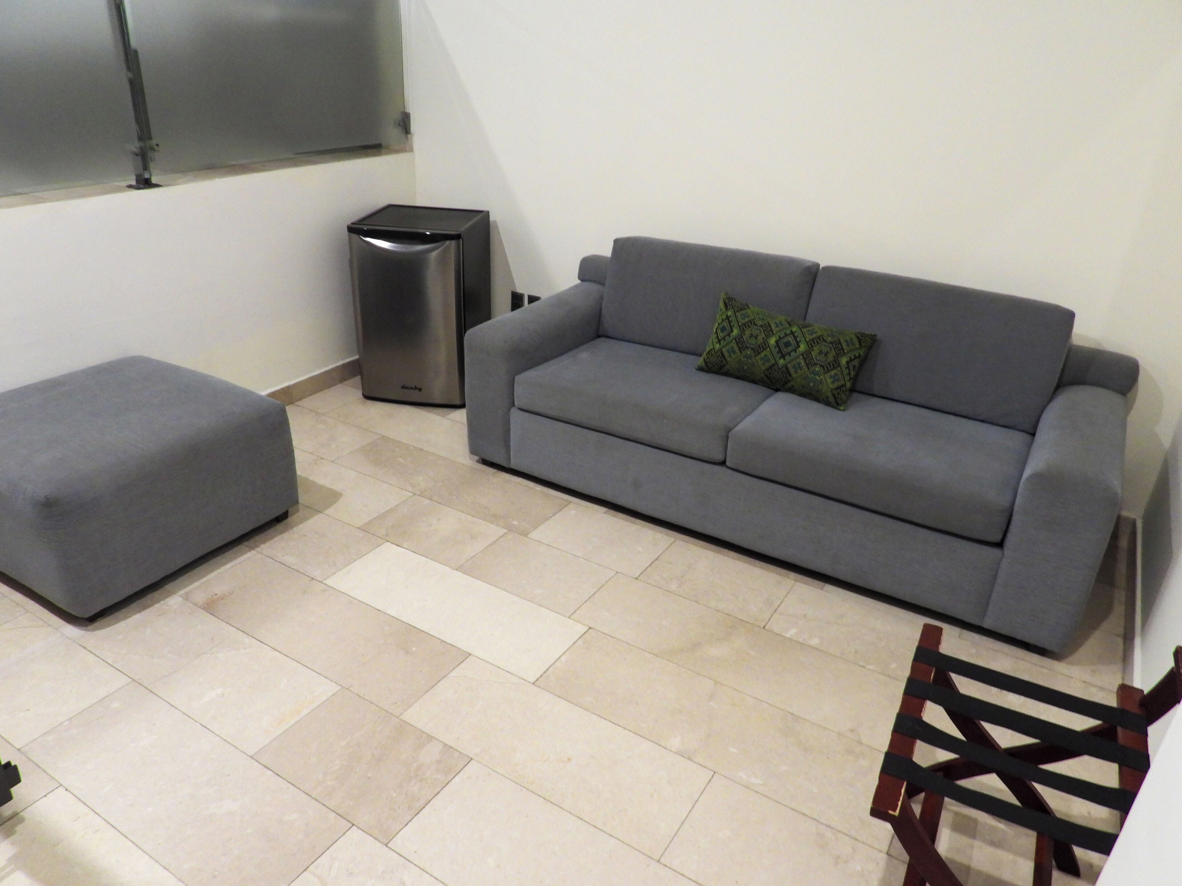 A living room with a grey sofa, a matching ottoman, a small trash bin, and a wooden chair.