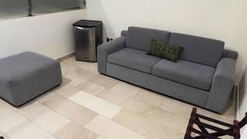 A living room with a grey sofa, a matching ottoman, a small trash bin, and a wooden chair.
