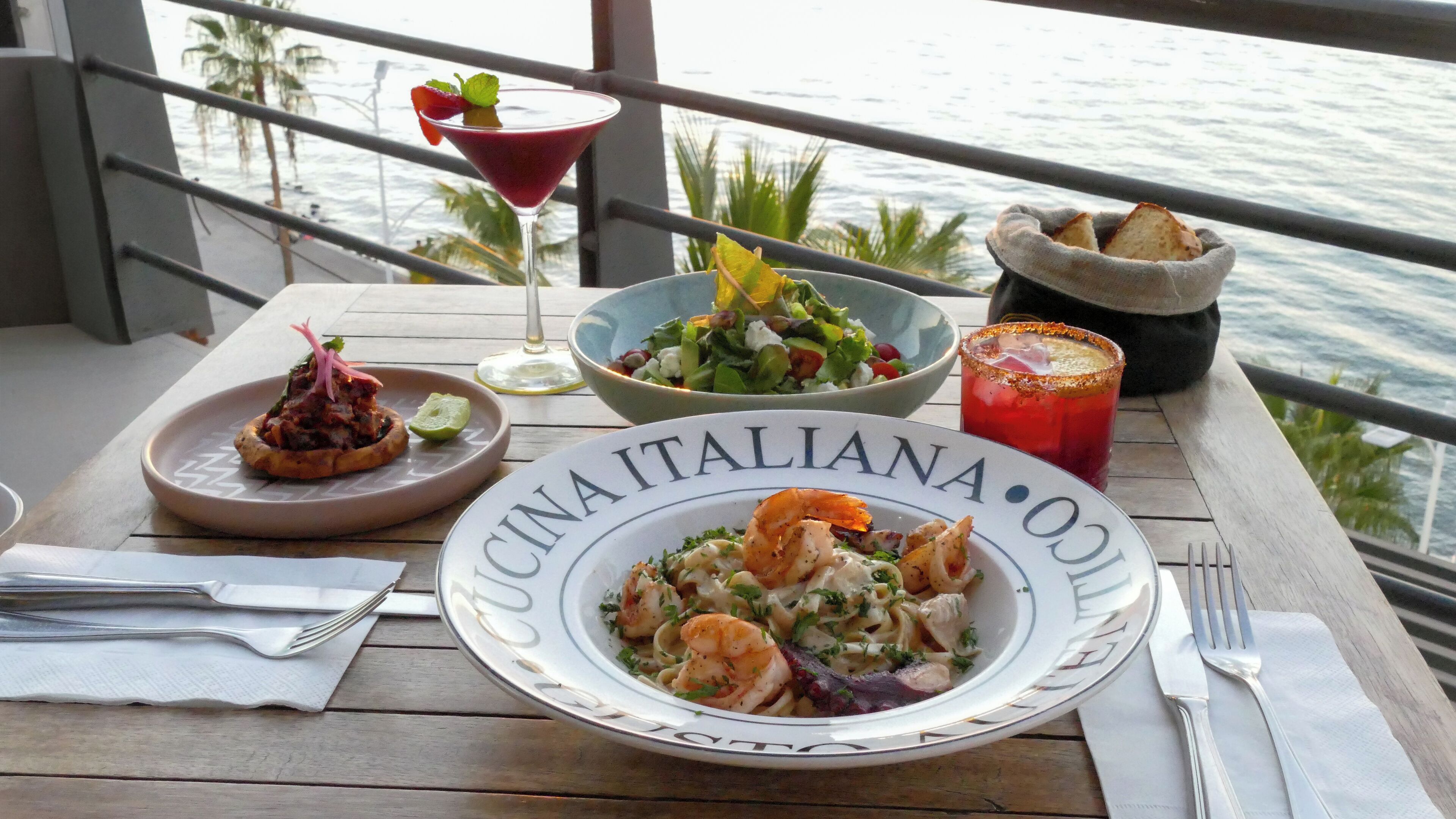 A dining table with a plate of pasta, a salad, and a cocktail on a balcony with a view of the sea.