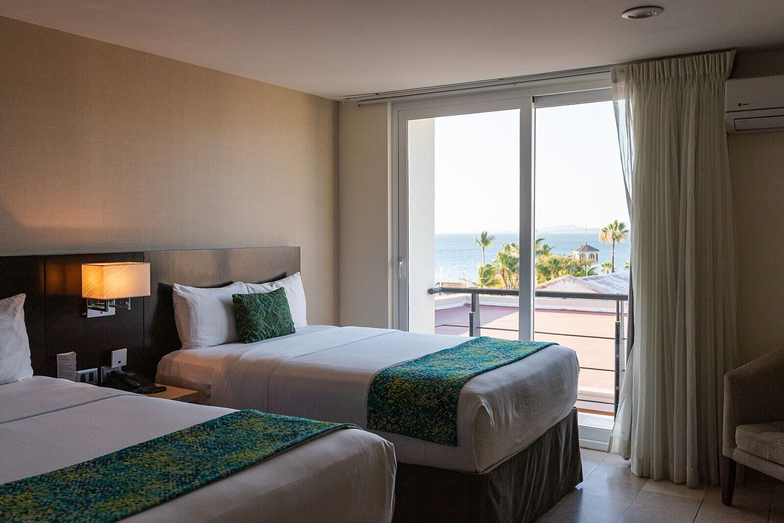 A hotel room with two beds, a nightstand, a lamp, a view of the ocean, and a balcony.