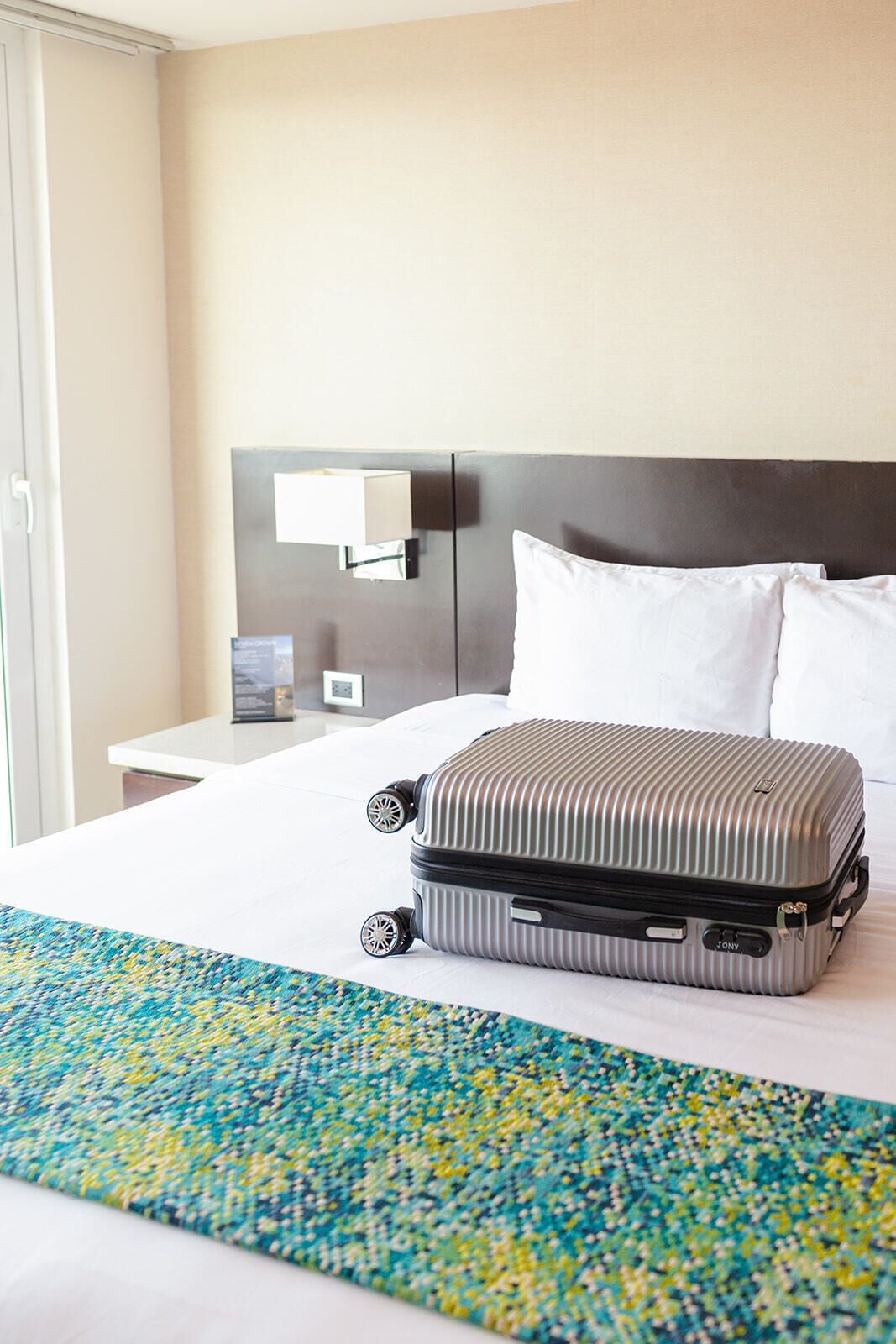 Quarto de hotel com uma cama, uma mala, uma mesa de cabeceira e uma luminária fixada na parede.