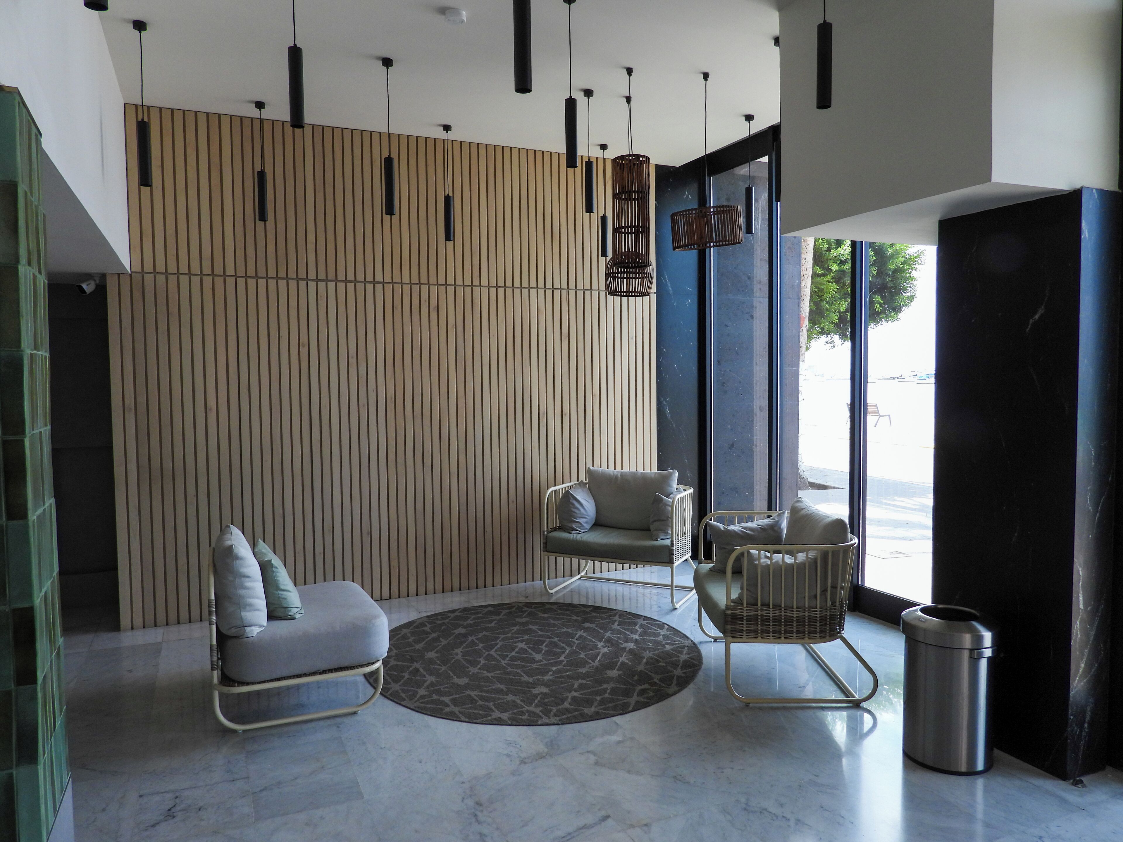 A modern interior with a wooden slat wall, two seating areas with cushions, a patterned rug, and hanging pendant lights.