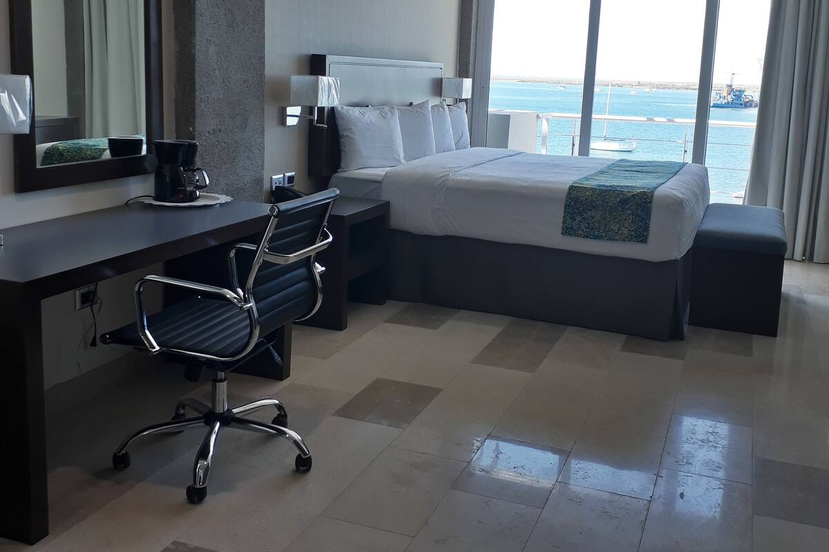 A hotel room with a large bed, a desk with a chair, a window with a view of the sea, and a mirror on the wall.