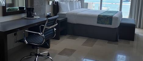 Una camera d'albergo con un letto grande, una scrivania con una sedia, una finestra vista mare e uno specchio appeso al muro.