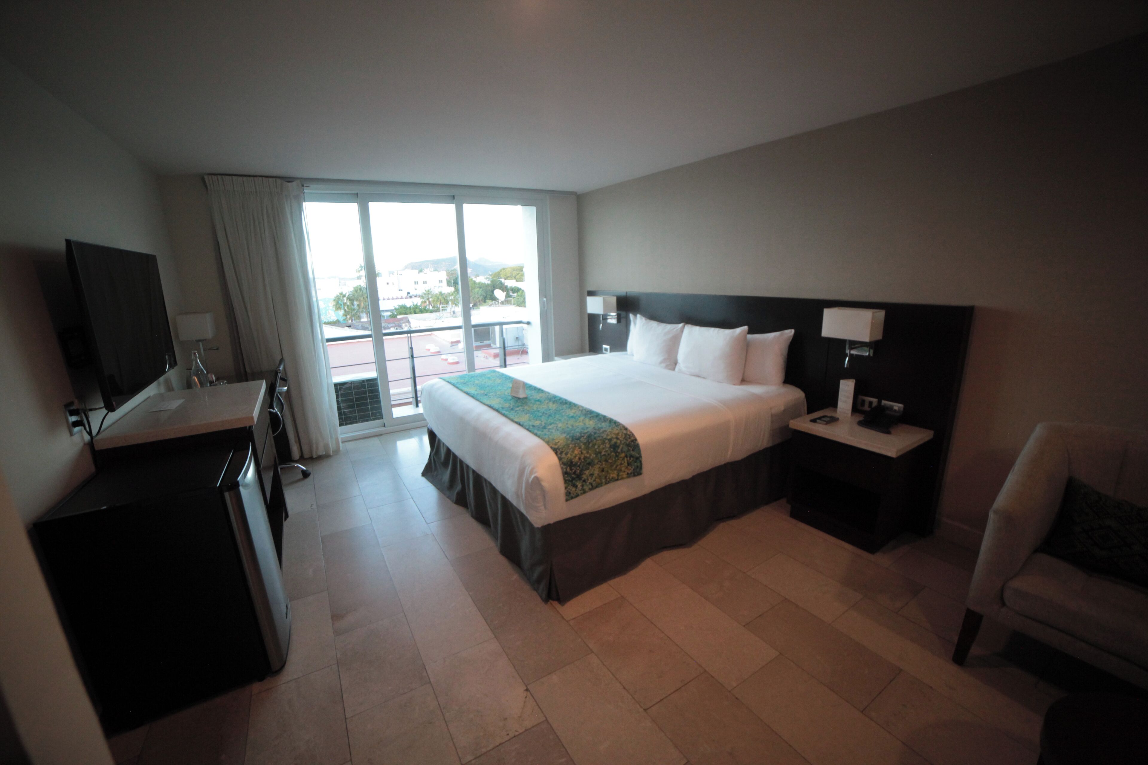 A hotel room with a large bed, a desk with a TV, a chair, and a view of the outdoors through a sliding door.