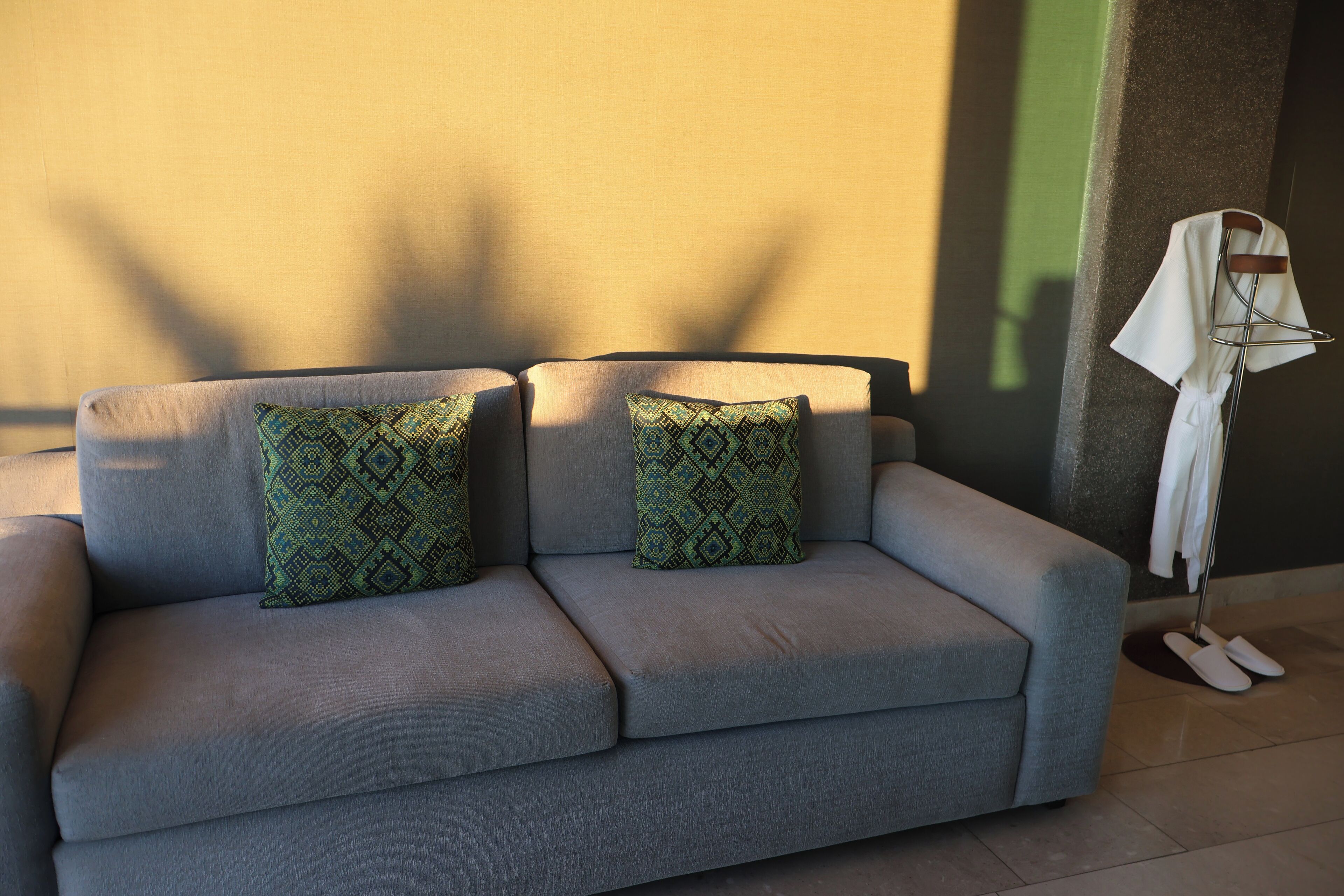 A grey sofa with patterned cushions, a white towel on a stand, and a wall with a shadow.