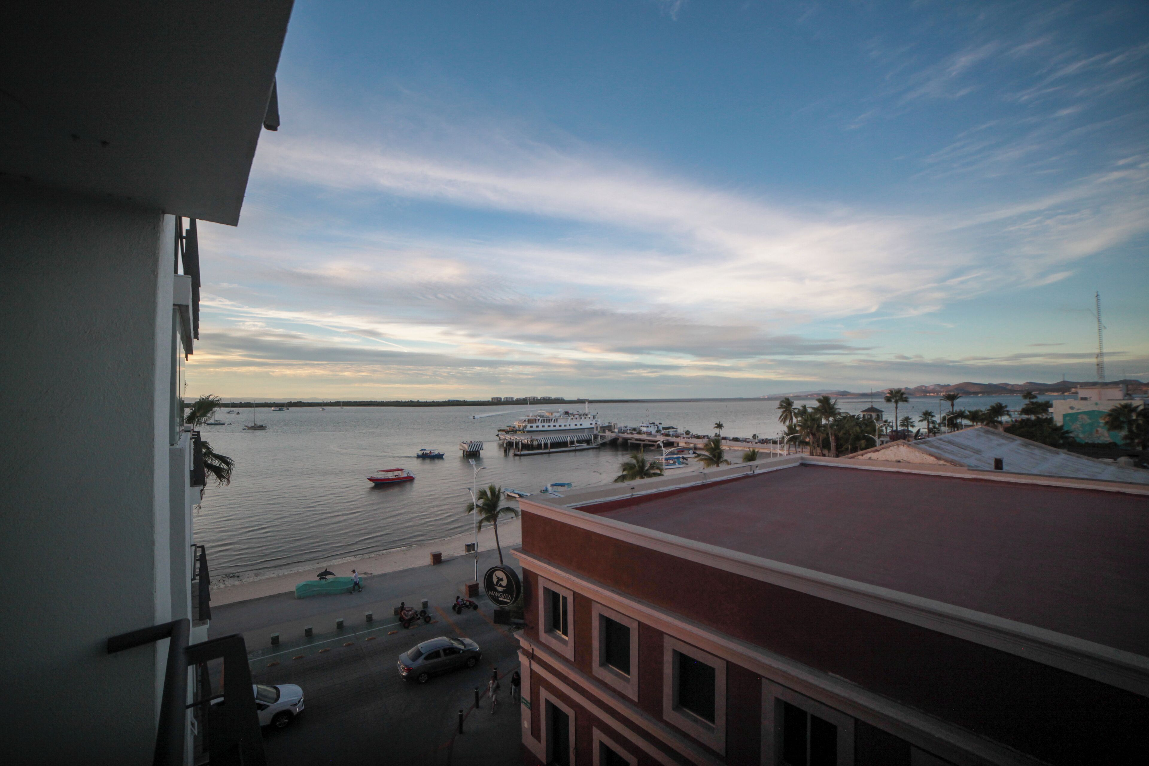 Vista costera desde un balcón con vista a un puerto deportivo con barcos, un muelle y una playa con palmeras.