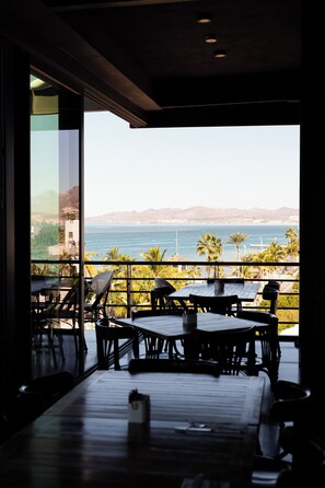 An indoor seating area with tables and chairs, a view of the sea, and a balcony with palm trees. - Seven Crown La Paz Malecon (La Paz)