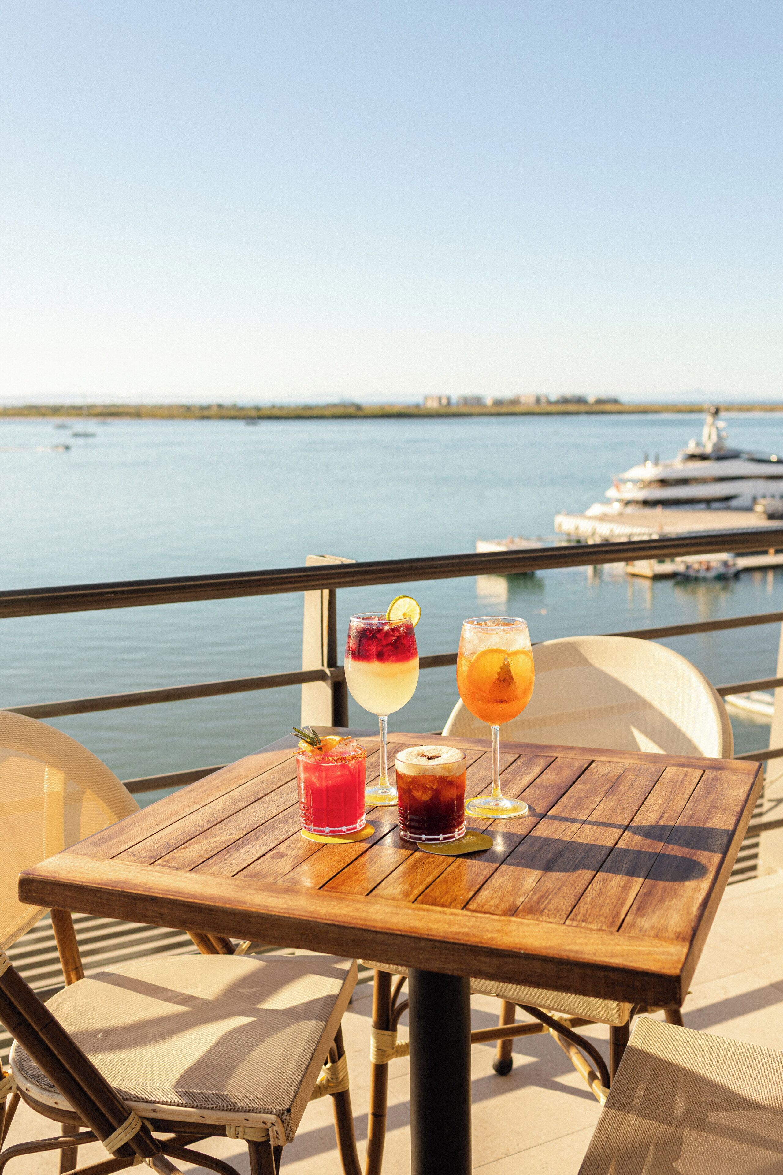 A wooden table with drinks and a view of the sea and a yacht.
