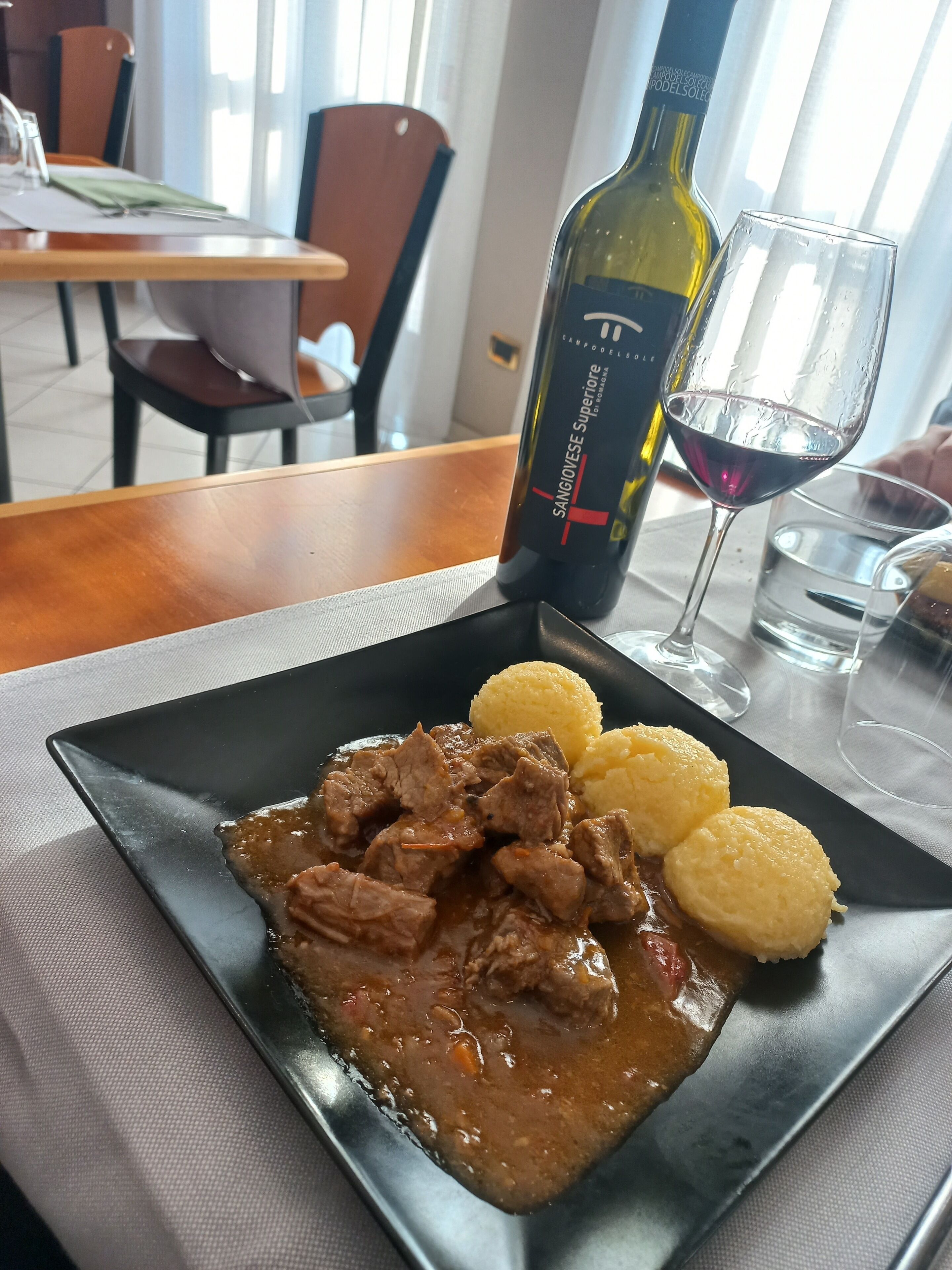 Ein Teller mit Rindereintopf und Knödeln auf einer schwarzen Platte, eine Weinflasche und ein Glas Rotwein auf einem Tisch in einem Restaurant.