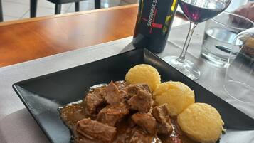 Ein Teller mit Rindereintopf und Knödeln auf einer schwarzen Platte, eine Weinflasche und ein Glas Rotwein auf einem Tisch in einem Restaurant.