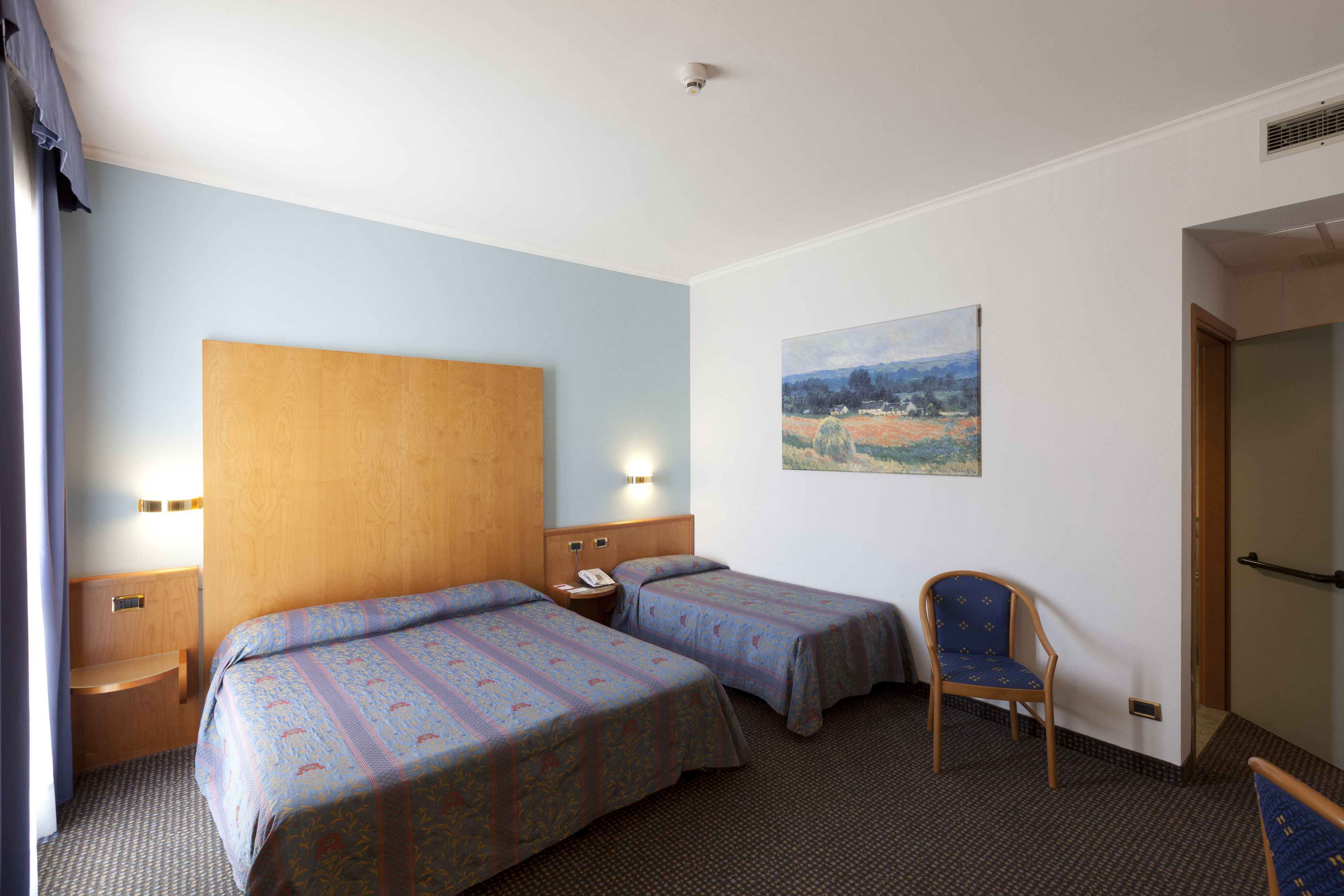 A hotel room with two beds, a wooden headboard, a painting on the wall, a chair, and a nightstand with a phone.
