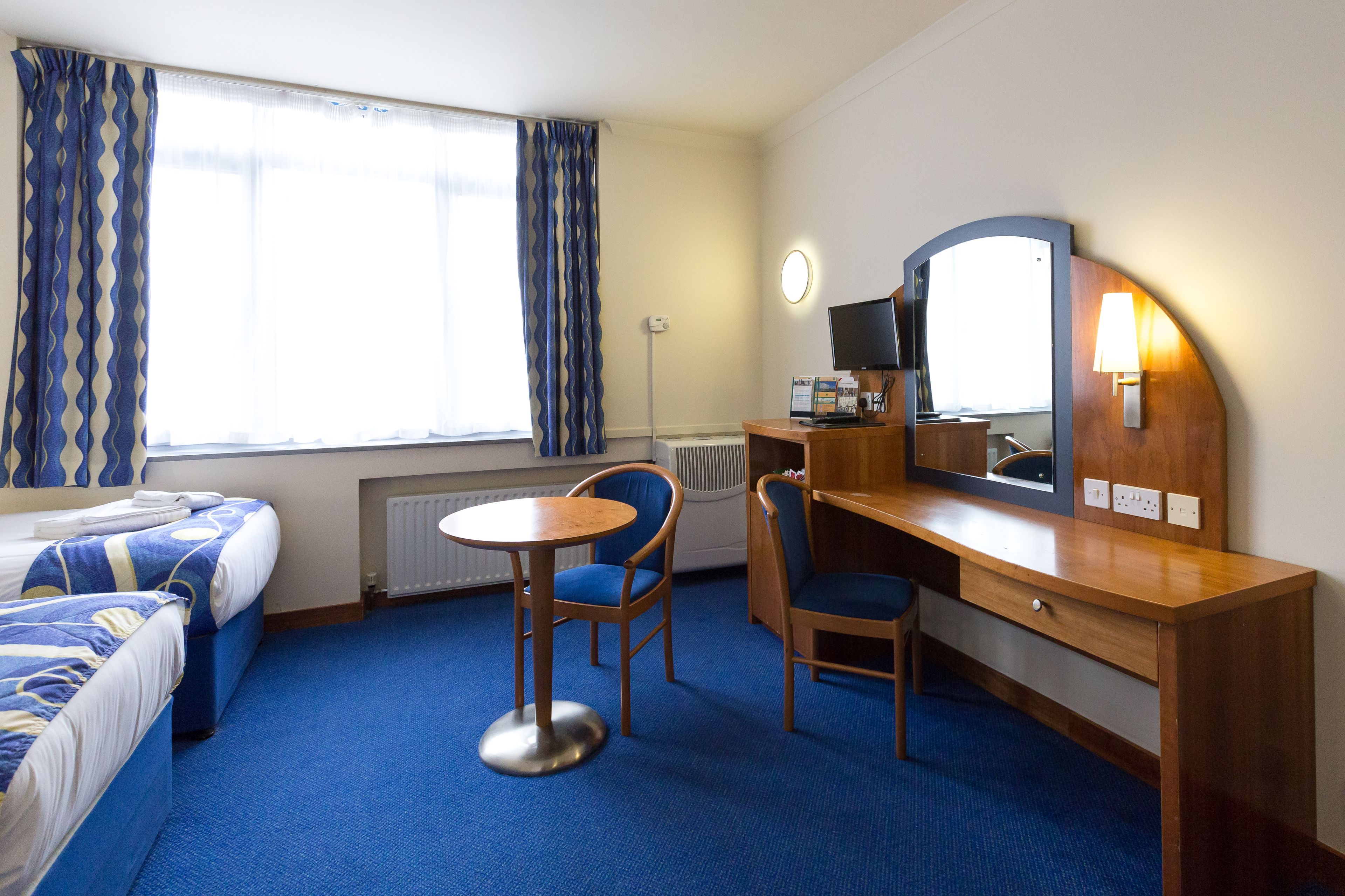 A hotel room with two beds, a desk with a chair, a round table, a mirror, and a window with curtains.