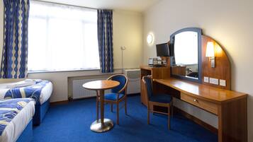 A hotel room with two beds, a desk with a chair, a round table, a mirror, and a window with curtains.