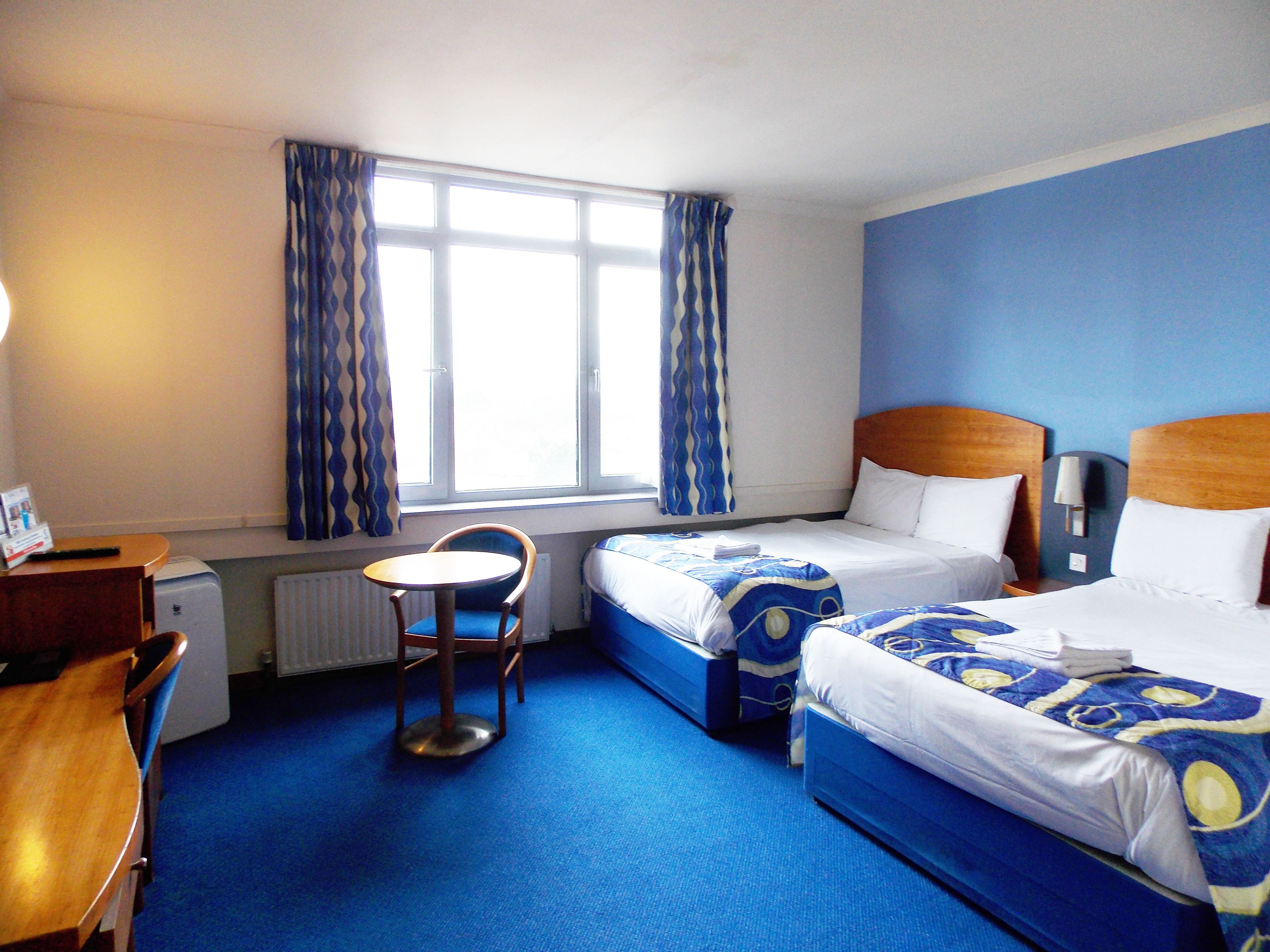 A hotel room with two beds, a small table, a chair, and a window with curtains.