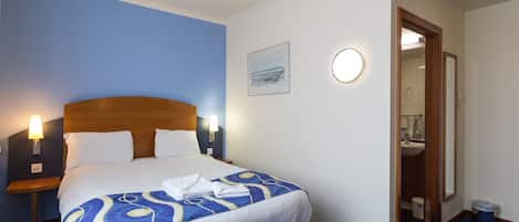 A hotel room with a bed, bedside lamps, a round table, a bathroom visible through an open door, and a framed picture on the wall.