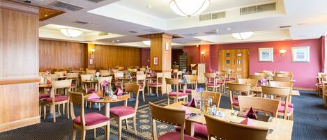 A hotel dining area with multiple tables and chairs, a patterned carpet, and a wooden reception desk.
