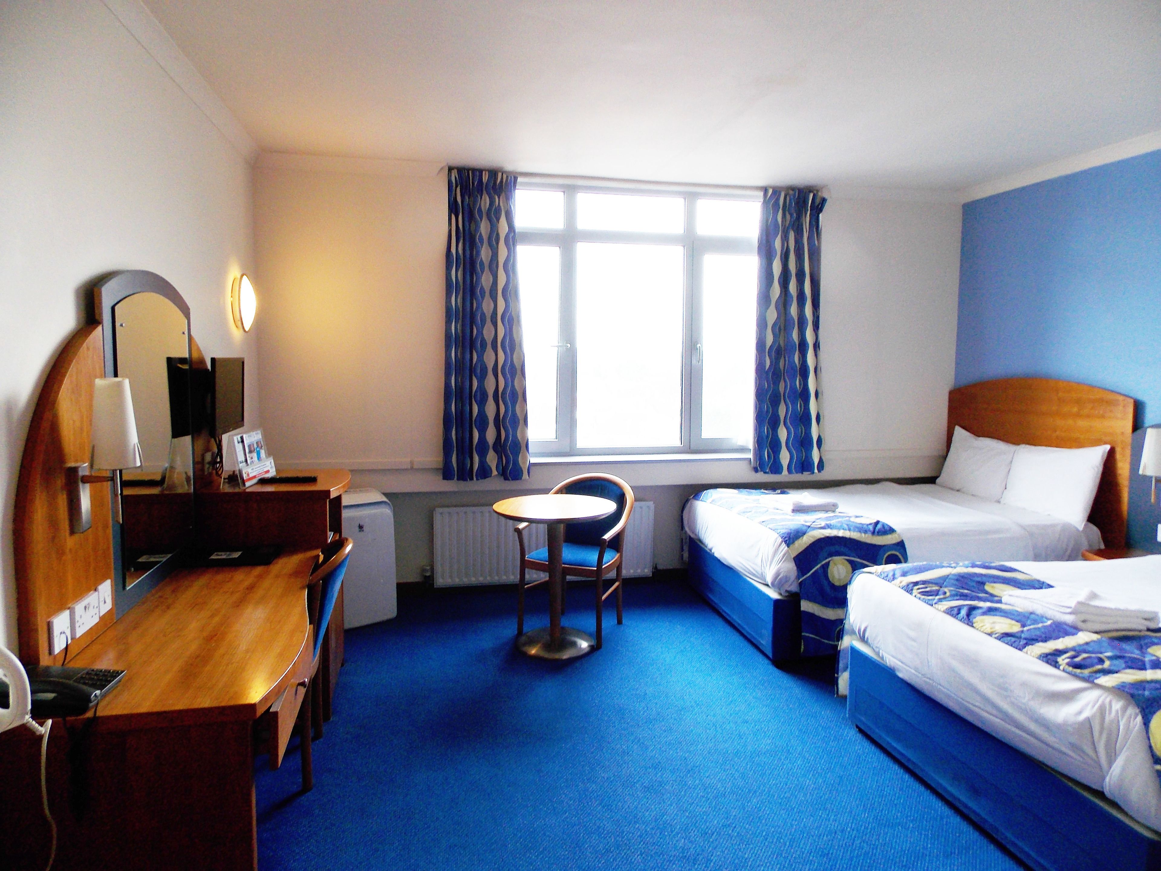 A hotel room with a wooden desk, a chair, a bed with blue bedding, and a window with blue curtains.