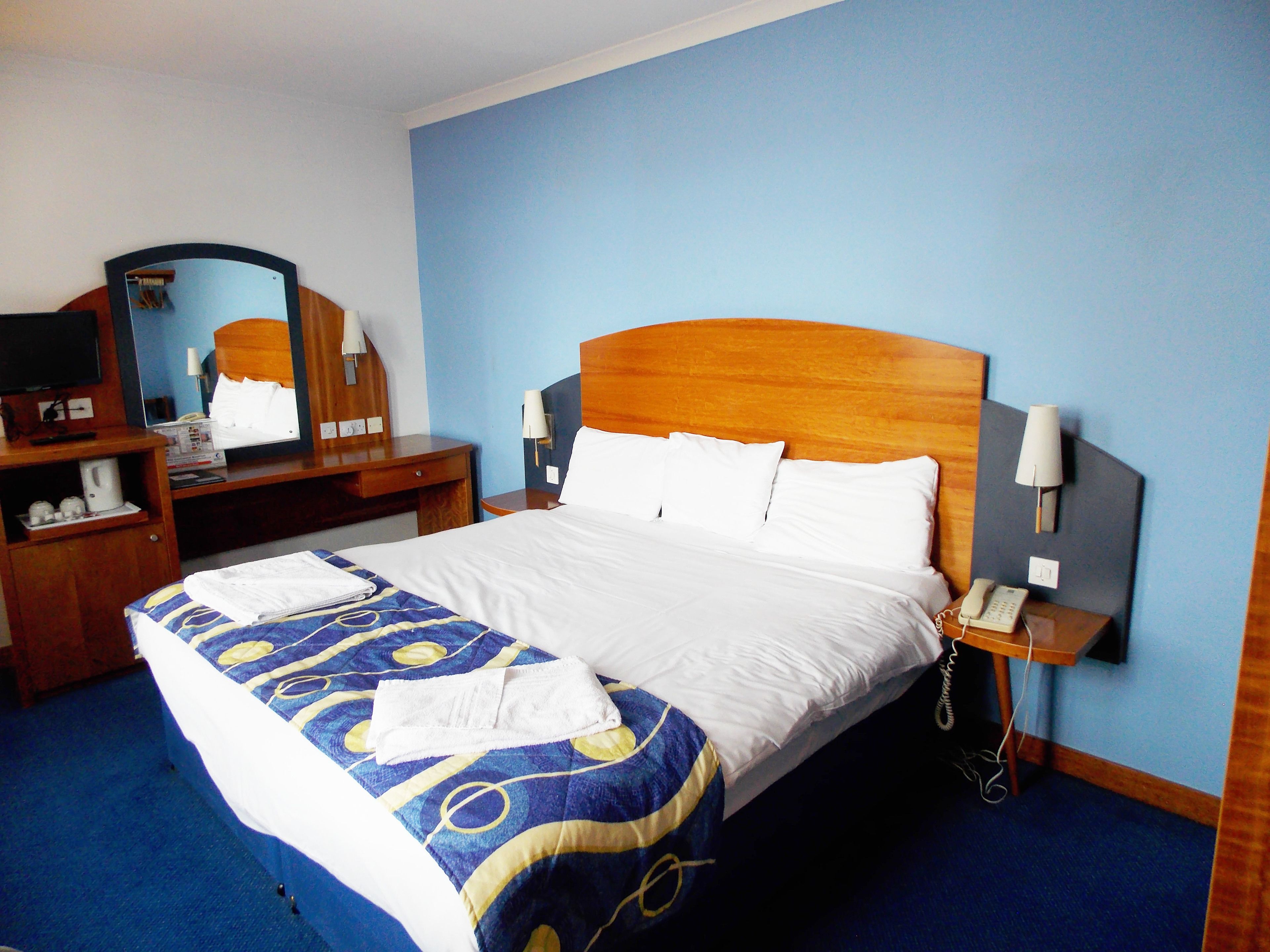 A hotel room with a bed, a desk with a telephone, a mirror, and a wall-mounted lamp.